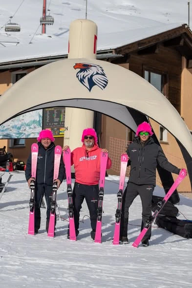 Three men wearing bright pink fuzzy hats standing under an Anavon branded tent at a ski resort, each holding a pair of bright pink Anavon skis.